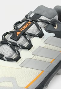 Γκρι αθλητικό παπούτσι με επάνω μέρος από δίχτυ, πορτοκαλί λεπτομέρειες, μαύρα κορδόνια και τρεις γκρι ρίγες στο πλάι για υποστήριξη και πρόσφυση.