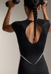 Fekete sportfelső, kivágott hátrészes dizájnnal és fehér díszítő vonalakkal. A modell súlyzókat tart, bemutatva a testhez simuló sziluettet.