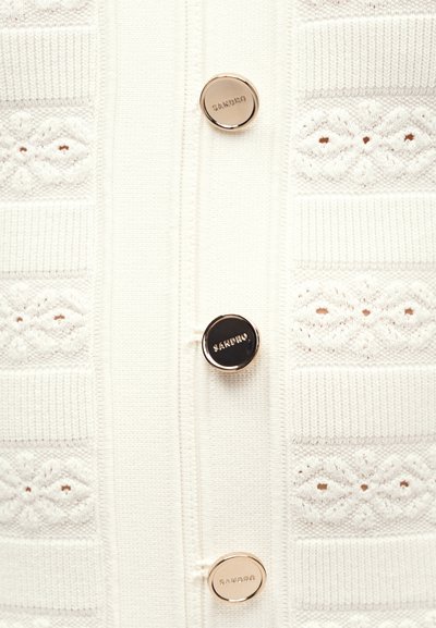 Λευκό πλεκτό ύφασμα με οριζόντια ανάγλυφα μοτίβα και τρία στρογγυλά χρυσά κουμπιά χαραγμένα με "SANDRO" κατά μήκος του κέντρου.