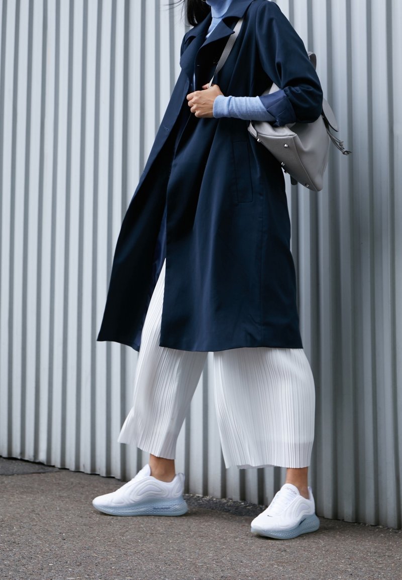Femme marchant à côté d'un mur en métal ondulé, vêtue d'un manteau bleu marine, d'un col roulé bleu clair, d'un pantalon plissé blanc, de baskets blanches, et portant un sac à main gris.