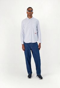 Šviesiai mėlyni dryžuoti marškiniai su krūtinės kišene ir raudonu logotipu, derinami su laisvai krentančiomis mėlynomis džinsais ir juodais sportbačiais. Dėvimos saulės akiniai.