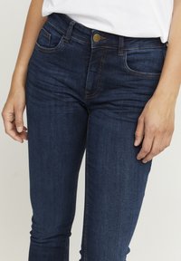 Temno modri jeans iz denima z oprijetim krojem, z zadrgo, gumbom in subtilnimi šivi na žepih.