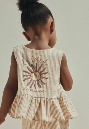 Kind mit dunklem lockigem Haar in einem gestreiften, beigen ärmellosen Oberteil mit Sonnenmotiv und französischem Text, steht mit dem Rücken zur Kamera vor einem schlichten Hintergrund.