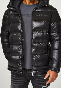 Sort dunjakke med en glansfuld finish, udstyret med lynlås, hætte og panelaccenter. Båret over en grå t-shirt med trykt logo.