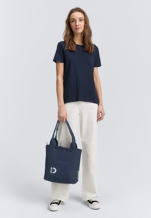 Mörkblå toteväska med texturerad tyg, dubbla handtag och en framficka med en ljusgrön logotyp. Bärs med en marinblå t-shirt och vita byxor.
