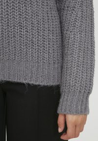 Grijze gebreide trui met een chunky textuur, geribde zoom en manchetten. Het garen lijkt fluffy, gecombineerd met slanke zwarte broek.