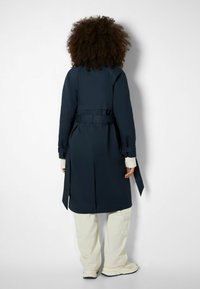 Trench coat blu navy con cintura in vita, maniche lunghe e spacco posteriore. Abbinato a pantaloni larghi color crema e sneakers.