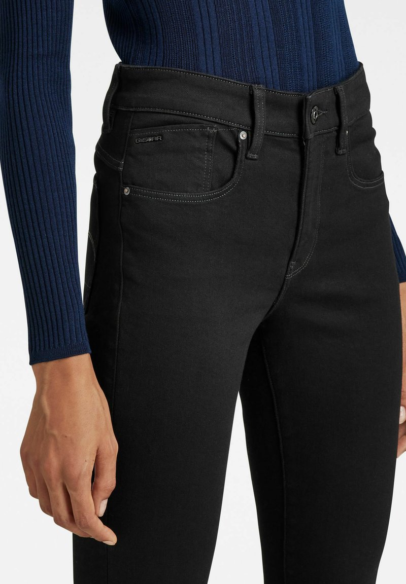 Schwarze Jeans aus Denim mit hohem Bund, Seitentaschen und kontrastierenden Nähten. Das Material hat eine glatte, leicht dehnbare Textur.