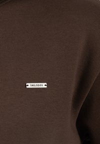 Brązowy sweter z teksturowanej tkaniny. Posiada małą metalową metkę z napisem "SMILODOX" w czarnym kolorze, przyszytą z przodu.