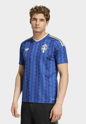Joven con camiseta azul del equipo nacional de fútbol de Suecia con detalles amarillos y logo de Adidas, de pie frente a un fondo liso.