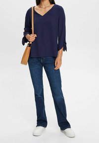 Blouse marine à col en V avec manches longues et accents noués aux poignets, associée à un jean bleu foncé et des baskets blanches. Sac à bandoulière marron inclus.