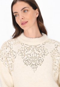 Krém színű kötött pulóver magas nyakkal, amelyet bonyolult arany flitter díszítések ékesítenek virágmintákban az elülső részen és a vállakon.