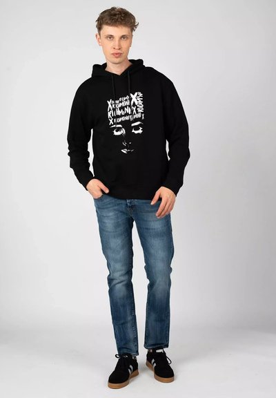 Felpa nera con cappuccio e design grafico che presenta un volto bianco, abbinata a jeans azzurri chiari e sneakers nere con dettagli bianchi.