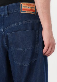 Jeans iz denima temno modre barve, z usnjeno oznako na hrbtni strani in žepom z našitkom za dekoracijo.