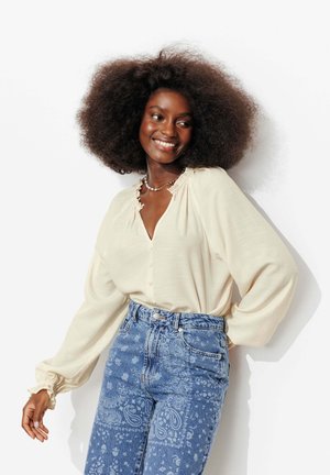 Femme souriante aux longs cheveux bouclés porte un chemisier crème à manches longues et un jean taille haute bleu à motif cachemire sur fond blanc.