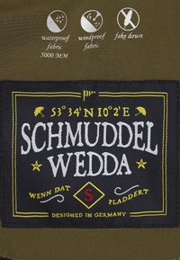 Grön vattentät tyg etikett med broderad text. Har symboler som indikerar vattentät, vindtät och konstgjord dunmaterial.