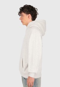 Mladý muž s kudrnatými tmavými vlasy, oblečený v světle šedé mikině a modrých džínách, stojící v profilu na jednotném pozadí.