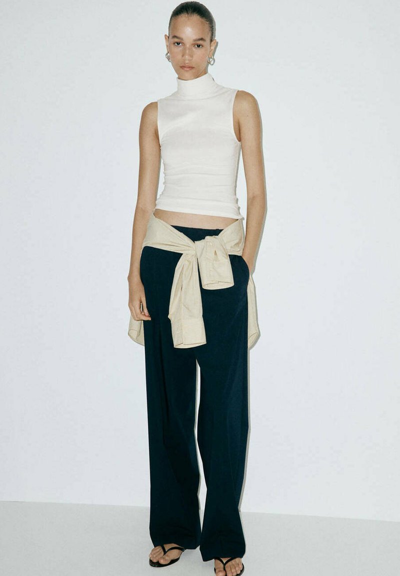 Femme aux cheveux attachés portant un col roulé blanc sans manches, un pantalon noir à jambes larges, une chemise beige nouée autour de la taille et des sandales noires.