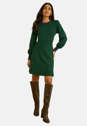 Grünes Strickkleid mit Puffärmeln, plissiertem Rock und pinkem, gewelltem Ausschnitt. Kombiniert mit braunen, hohen Stiefeln mit Absatz.