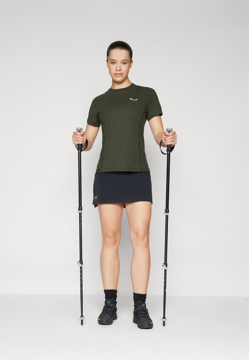 Krótki rękaw oliwkowej zielonej koszuli i czarnej spódnicy, sparowane z kijami trekkingowymi i butami do wędrówek, na neutralnym tle.