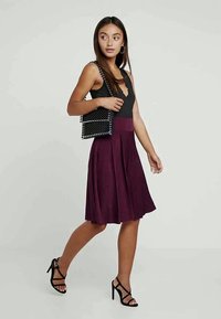 Mulher a caminhar, vestindo uma blusa preta sem mangas, uma saia roxa, sandálias de salto pretas e a carregar uma bolsa de ombro preta com contas.