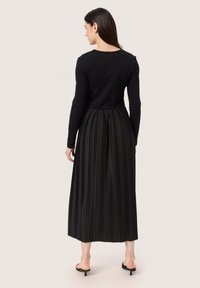 Black long-sleeve top connected to a pleated black skirt. Features a textured, flowing design with a fitted bodice and mid-calf length.