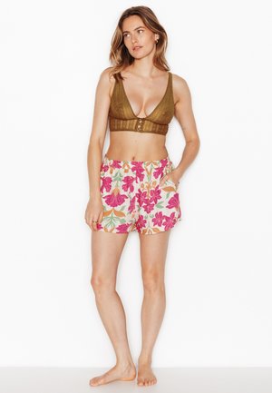 Femme debout pieds nus portant un bralette en dentelle marron et un short blanc avec un imprimé floral rose et orange, une main dans la poche, regardant sur le côté.
