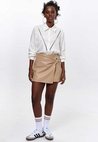 Witte blouse met lange mouwen en ingewikkelde details, gecombineerd met een beige faux leren wikkeltop. Model draagt witte sokken met gouden strepen en sneakers.