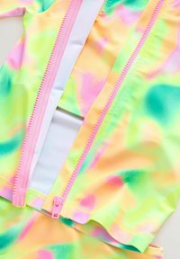 Maillot de bain coloré avec un motif ondulé en vert pastel, rose et jaune. Comprend une fermeture éclair rose et un tissu lisse et extensible.