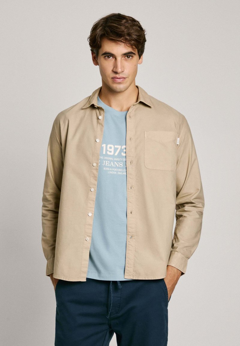 Camicia beige a maniche lunghe in cotone, con chiusura a bottoni e taschino sul petto, indossata sopra una maglietta grafica azzurro chiaro, abbinata a pantaloni blu navy.