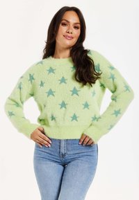 Green fuzzy sweater with a star pattern in light blue. Features long sleeves and a ribbed hemline, worn with light blue jeans.