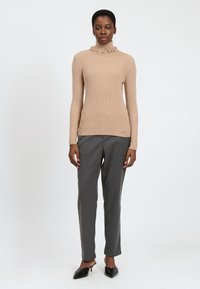 Donna in piedi contro uno sfondo bianco, indossa un maglione beige a coste con collo alto, pantaloni grigi dritti e mule nere con punta appuntita e tacco.