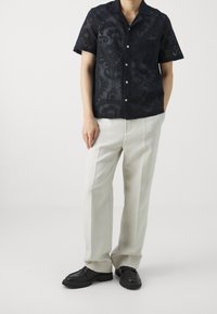Modello maschile che indossa una camicia nera trasparente a maniche corte con bottoni, pantaloni bianchi plissettati e mocassini neri su uno sfondo neutro.
