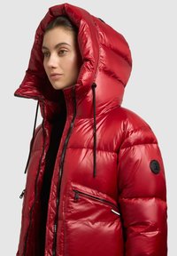 Sarkana pūķa jaka ar spīdīgu virsmu, lielu kapuci, rāvējslēdzēja priekšējām kabatām un kapuces regulējumiem ar auklu. Visa jaka ir veidota ar ķīļotu dizainu.