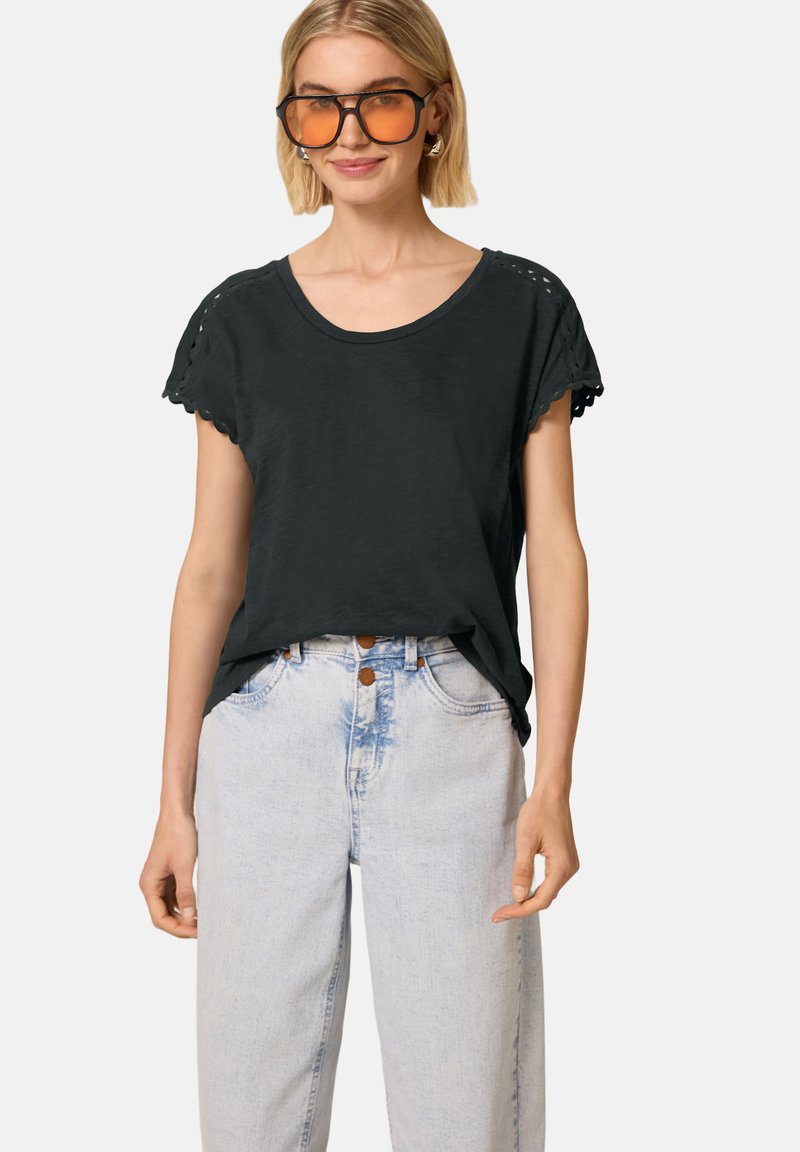 Femme portant de grandes lunettes de soleil teintées orange, un chemisier noir à manches courtes avec des détails en dentelle et un jean en denim bleu clair taille haute.