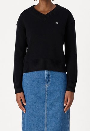 Czarny sweter z długim rękawem i dekoltem w serek z małym logo "CK", zestawiony z niebieską, wysoką spódnicą z denimu.