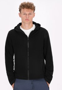 Črna zip-up kapuca iz mehkega materiala, z zadrgo spredaj, dvema stranskima žepoma in kapuco z vrvico; minimalistična zasnova.