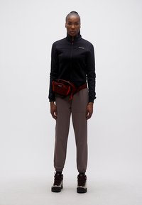 Γυναίκα που στέκεται όρθια φορώντας μια μαύρη ζακέτα με φερμουάρ, γκρι παντελόνι, μπότες πεζοπορίας και μια κόκκινη τσάντα μέσης σε απλό φόντο.