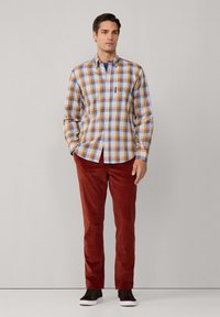 Camisa de cuadros en azul, naranja y blanco; combinada con pantalones de pana rojos. El modelo lleva zapatillas oscuras; diseño minimalista, sin patrones en los pantalones.