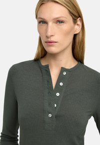 Długi rękaw, prążkowany top w ciemnozielonym kolorze z dekoltem henley, z kilkoma białymi guzikami i teksturowanym wzorem materiału.