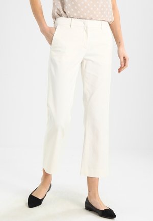 Kobieta w kremowych, krótkich spodniach, beżowej bluzce w grochy włożonej do środka oraz czarnych, spiczastych balerinach, stojąca na jednolitym tle.