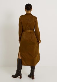 Vestido camisero largo de color marrón, acabado satinado, con cintura ceñida, puños abotonados, slit laterales, combinado con botas de tacón negras hasta la rodilla.