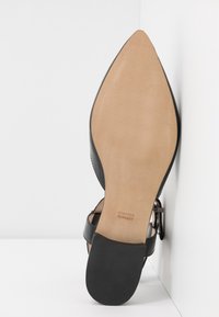 Semelle d'une sandale noire à bout pointu avec un dessous en cuir beige lisse et un talon noir texturé, nom de la marque visible près de l'arche.