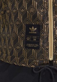 Bunda s černým a zlatým vzorem, která má zipové zapínání, texturovanou látku a viditelné logo Adidas s pokyny pro údržbu a velikost.