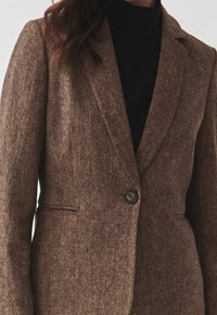 Ruskea teksturoitu bleiseri, jossa on yksittäinen nappisuljin, notched-kaulukset ja kapea istuvuus. Päällä musta pooloneule.