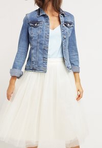 Chaqueta de mezclilla de lavado claro con dos bolsillos en el pecho, cierre de botones y puños doblados, combinada con una falda de tul blanca. Textura suave, diseño informal.