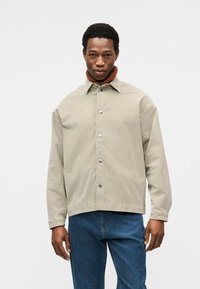 Veste en velours côtelé beige clair avec un col robuste, des boutons argentés et un col roulé côtelé marron. Associée à un jean bleu.