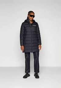 Sort polstret lang frakke med lynlås og hætte. Har et logo på brystet, parret med mørke bukser og sporty sorte sko.