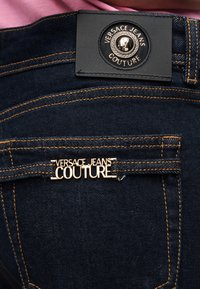 Τσέπη σκούρου μπλε τζιν με χρυσό έμβλημα "Versace Jeans Couture" και μαύρη δερμάτινη ετικέτα ραμμένη πάνω από την τσέπη.