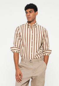 Gestreiftes Hemd in Beige- und Brauntönen; mit langen Ärmeln, Kragen und einem kleinen Logo auf der Brusttasche. Hose in Hellbeige.